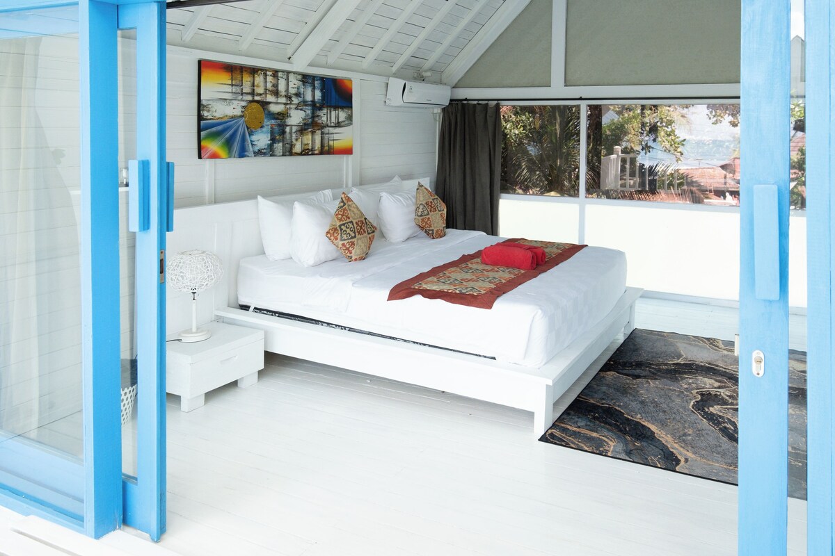 A modern bedroom with a white bed, patterned pillows, a red throw, and a large window with a view.