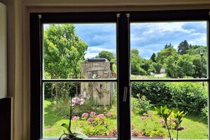 A room with a large window offering a view of a garden with various plants and a wooden structure. - Holiday apartment with 2 bedrooms (Mirow\/ Ortsteil Babke)