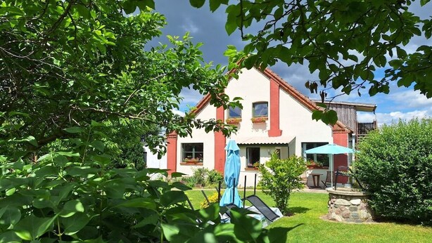 Una casa de dos pisos amb un teulat vermell, un balcó i un jardí amb un trampolí i mobles d'exterior.