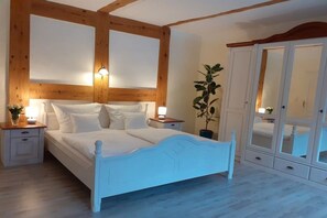 Ein Schlafzimmer mit einem weißen Bett, Holzbalkendecke, Nachttischen mit Lampen, einer Topfpflanze und einem Kleiderschrank mit Glastüren.