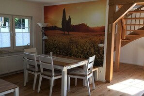 A dining area with a wooden table and white chairs, a mural of a field with a tree, and a staircase. - Holiday home at Eichenfleck (Seedorf)