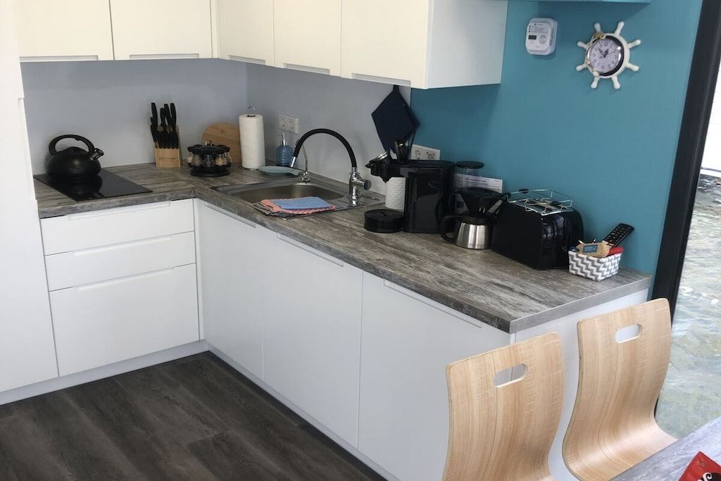 Een compacte keuken met witte kastjes, een gootsteen, een waterkoker, een koffiezetapparaat en een klok aan de muur.