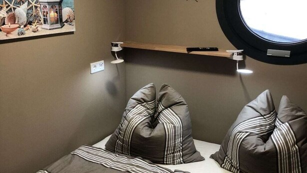 Un dormitorio con cama, ropa de cama a rayas, un estante en la pared, un ventanal redondo y un adorno mural.
