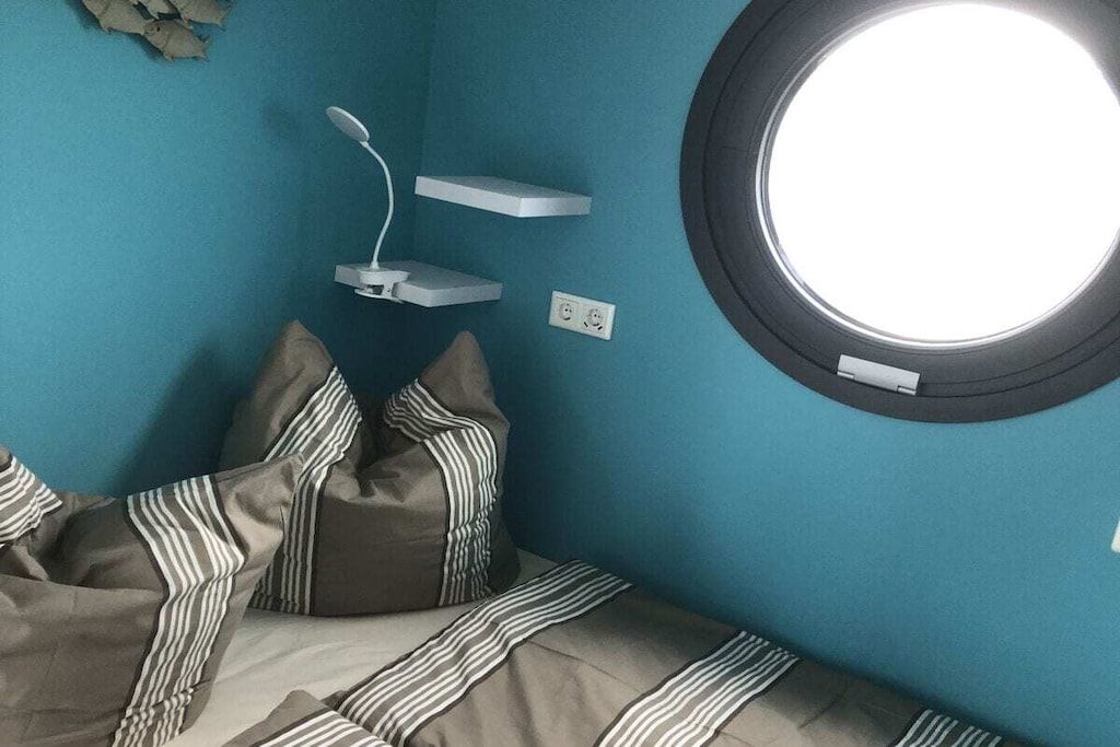 Ein Zimmer mit einem Bett, gestreifte Kissen, einem runden Fenster und einer weißen Ladestation.