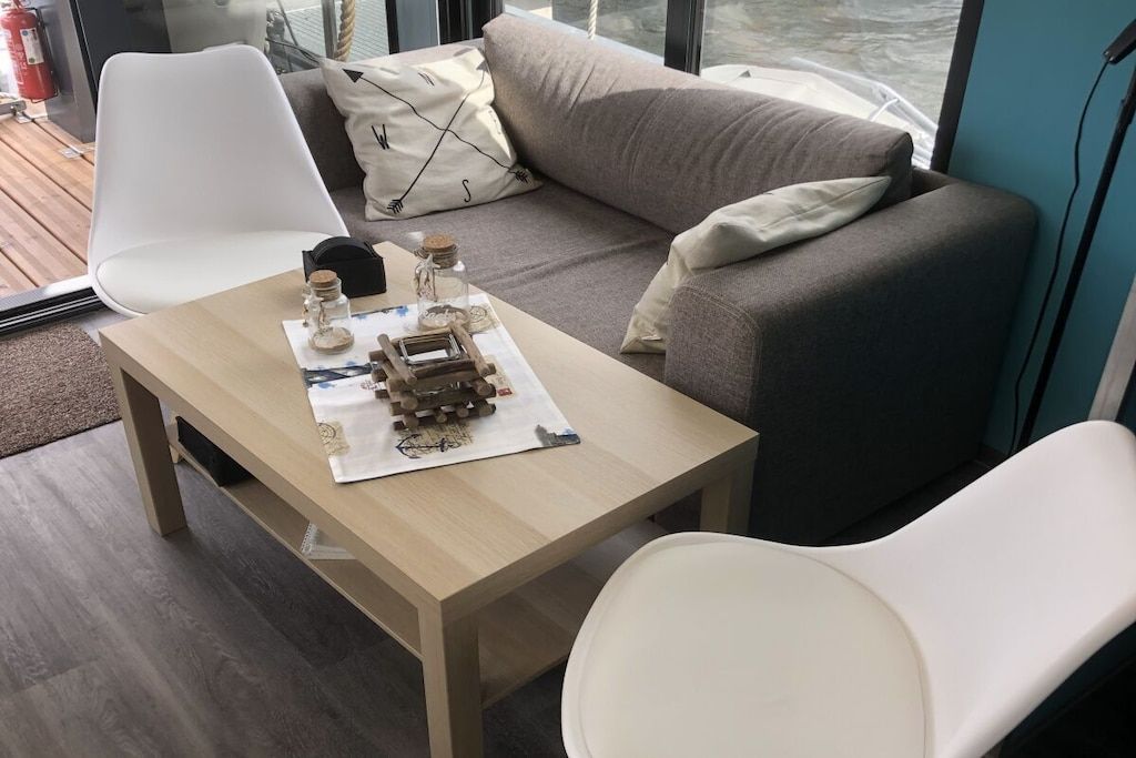 Ein modernes Wohnzimmer mit einer Couch, zwei weißen Sesseln, einem Holztisch mit einer Modelleisenbahn und einem Fenster mit Blick auf das Meer.