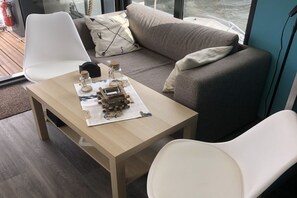 Ein modernes Wohnzimmer mit einer Couch, zwei weißen Sesseln, einem Holztisch mit einer Modelleisenbahn und einem Fenster mit Blick auf das Meer.
