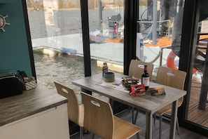 A table with chairs, a bottle, and a bag of snacks in a room with large windows overlooking a dock area. - myHolidayBoat Krimnicksee (Königs Wusterhausen)