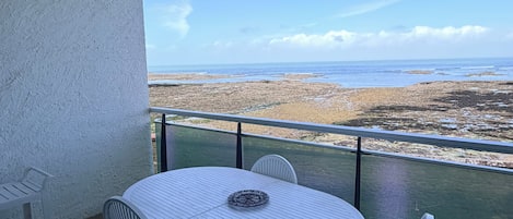 A balcony with a white table and chairs, overlooking a coastal landscape.