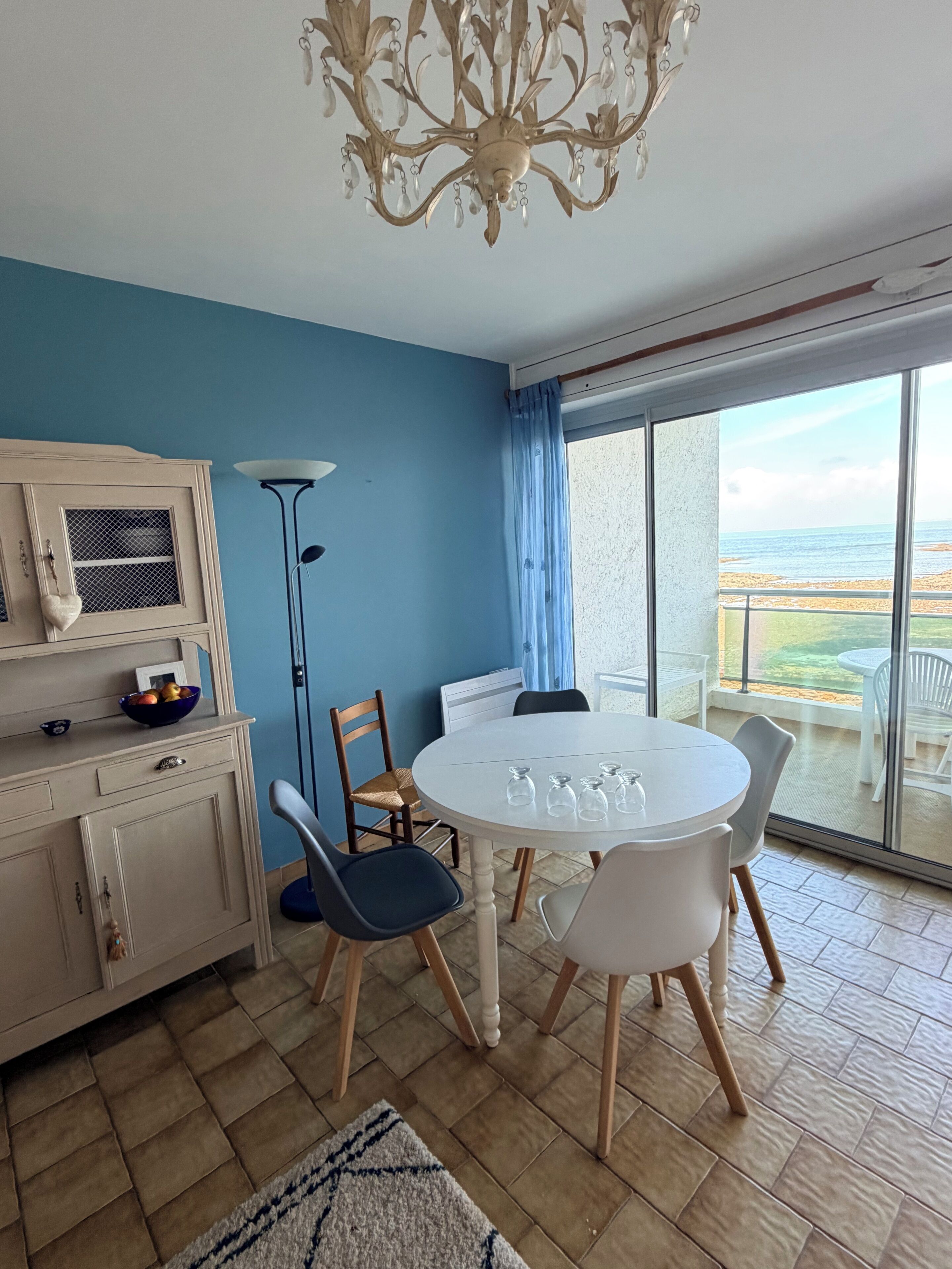Quarto com mesa de jantar, cadeiras, lustre, armário e vista para o mar através de grandes janelas.
