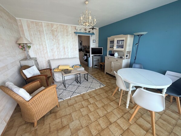 Un salon comprenant un canapé, des fauteuils, une table basse, une table à manger avec des chaises, un lustre et un coin cuisine.
