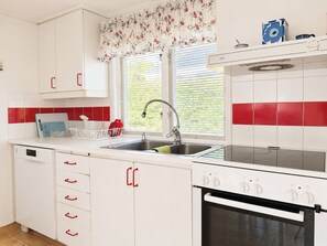 A kitchen with white cabinets, a double sink, a stove, and a window with blinds. - Jennys Pärla by Interhome (Haverdal)