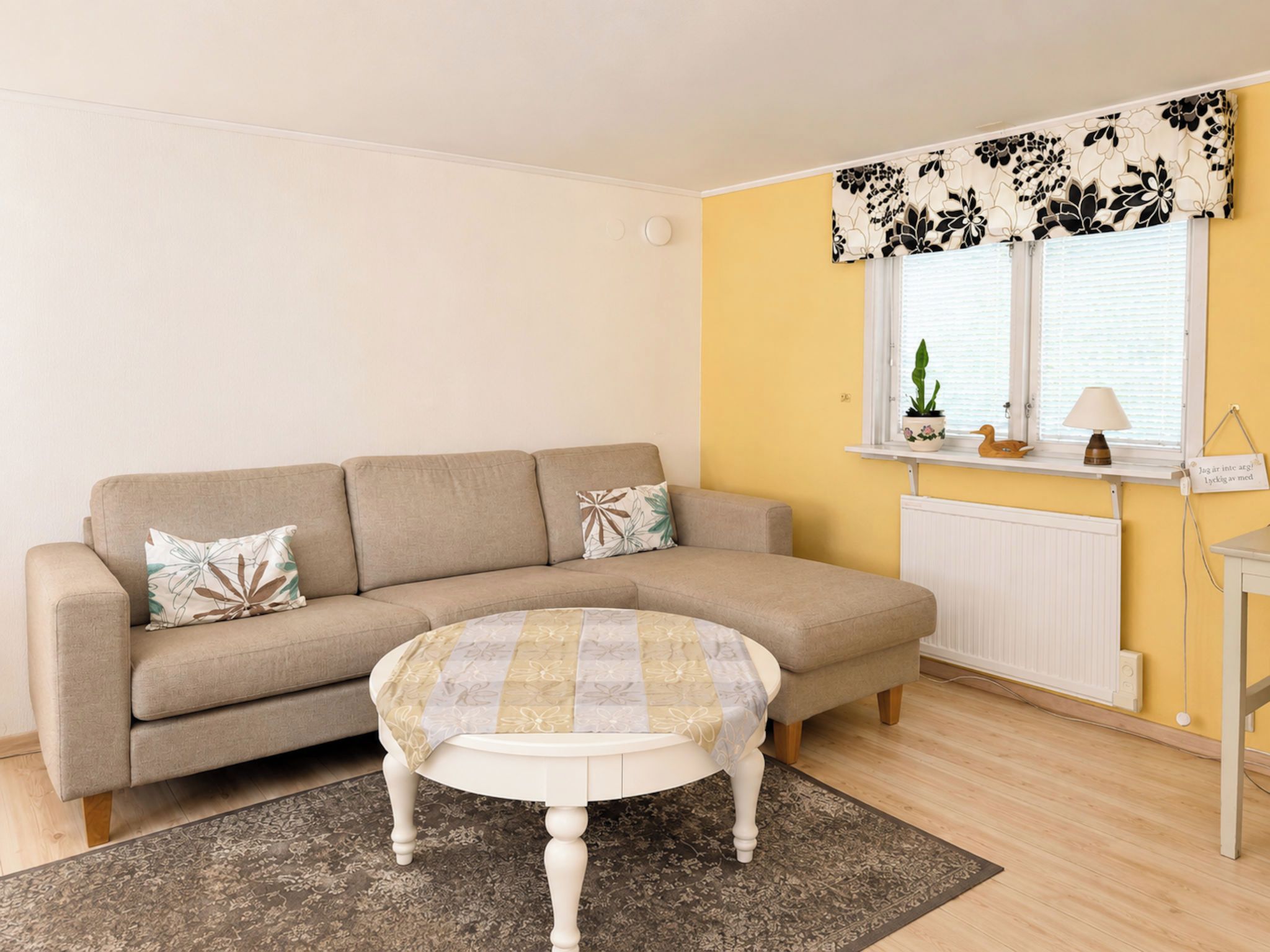 A living room with a sofa, a round coffee table, a rug, a window with blinds, a radiator, and a small shelf with a plant and a lamp.