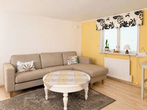 A living room with a sofa, a round coffee table, a rug, a window with blinds, a radiator, and a small shelf with a plant and a lamp. - Jennys Pärla by Interhome (Haverdal)
