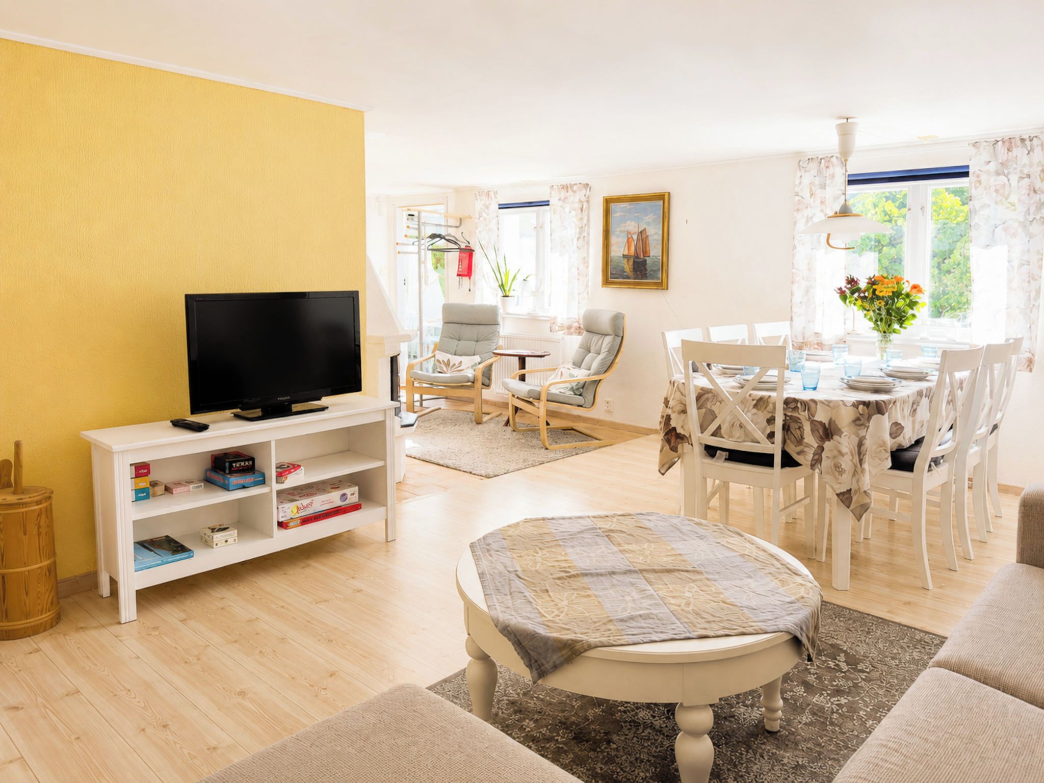 Una sala de estar con televisor, estantería, mesa de comedor con sillas y ventana con cortinas.