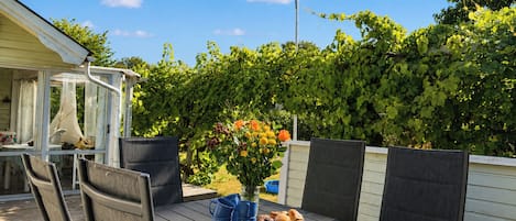 An outdoor patio with a table set for dining, surrounded by chairs, and a garden with a fence and greenery in the background.