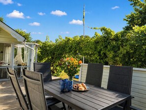 An outdoor patio with a table set for dining, surrounded by chairs, and a garden with a fence and greenery in the background. - Jennys Pärla by Interhome (Haverdal)