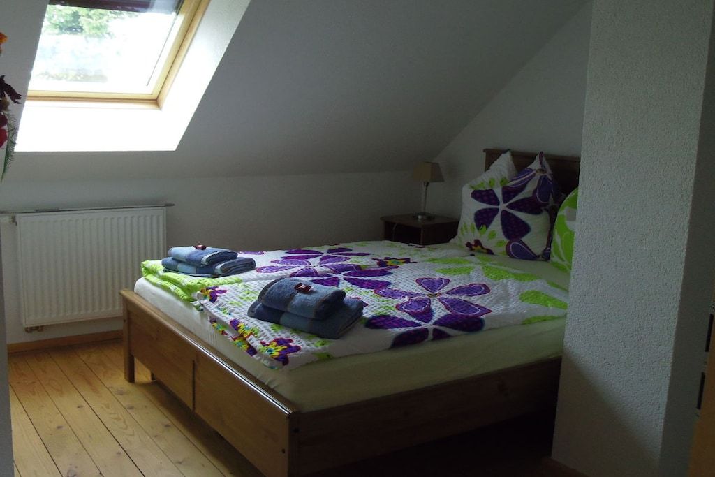 Ein Einzelbett mit floralem Bettbezug, ein Nachttisch, eine Lampe und ein Fenster mit Blick auf Grünfläche.