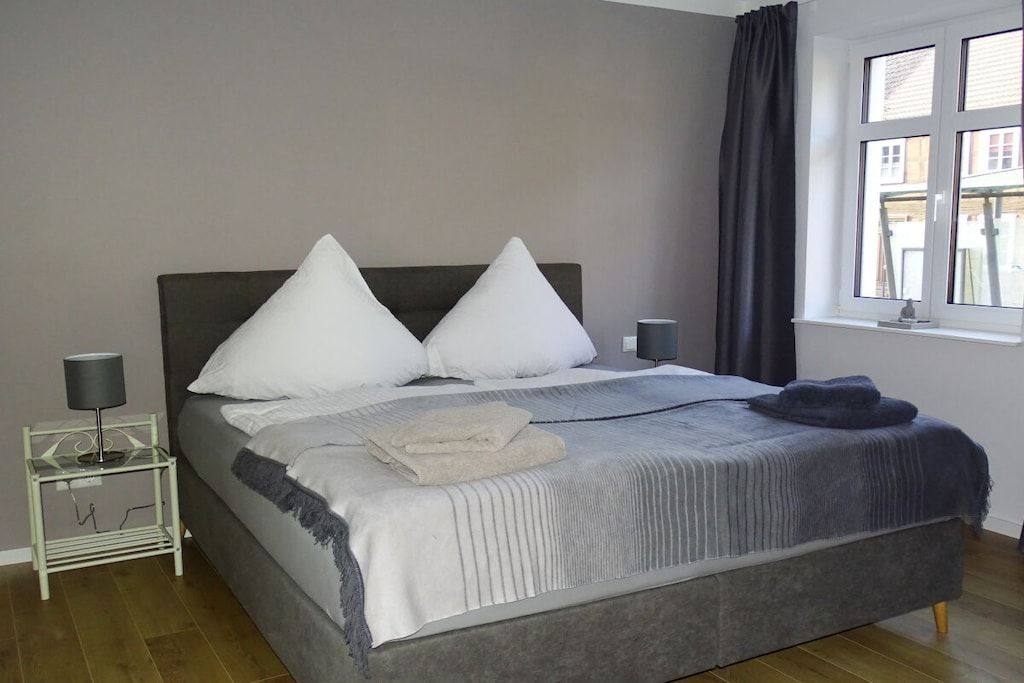 Un dormitorio con cama, almohadas, una mesita de noche con lámpara, una ventana con cortinas y vistas a edificios.