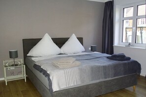 Ein Schlafzimmer mit Bett, Kissen, einem Nachttisch mit Lampe, einem Fenster mit Vorhängen und Blick auf Gebäude.