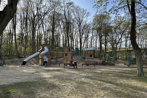 A playground with a slide and swings, surrounded by trees and a wooden structure. - "the house by the lake" (Malchow)