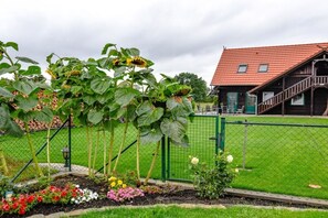 Ein von einer Hecke umgebener Garten mit verschiedenen Blumen und Sonnenblumen, ein Holzgebäude mit rotem Dach und ein grüner Zaun.