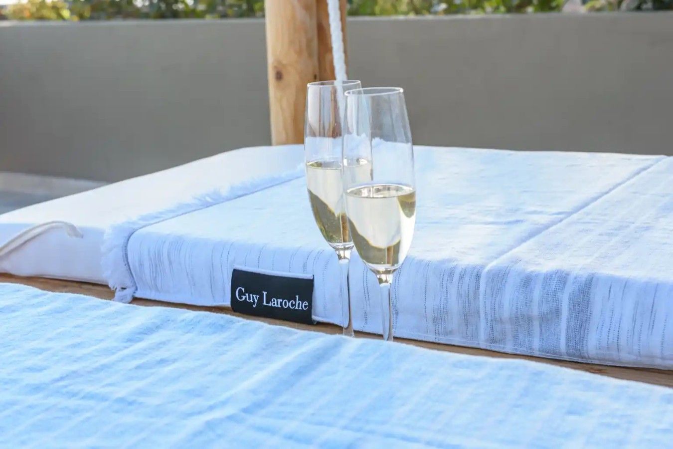 Cama de hotel com etiqueta de nome indicando 'Guy Laroche' e dois copos de vinho branco sobre a cama.