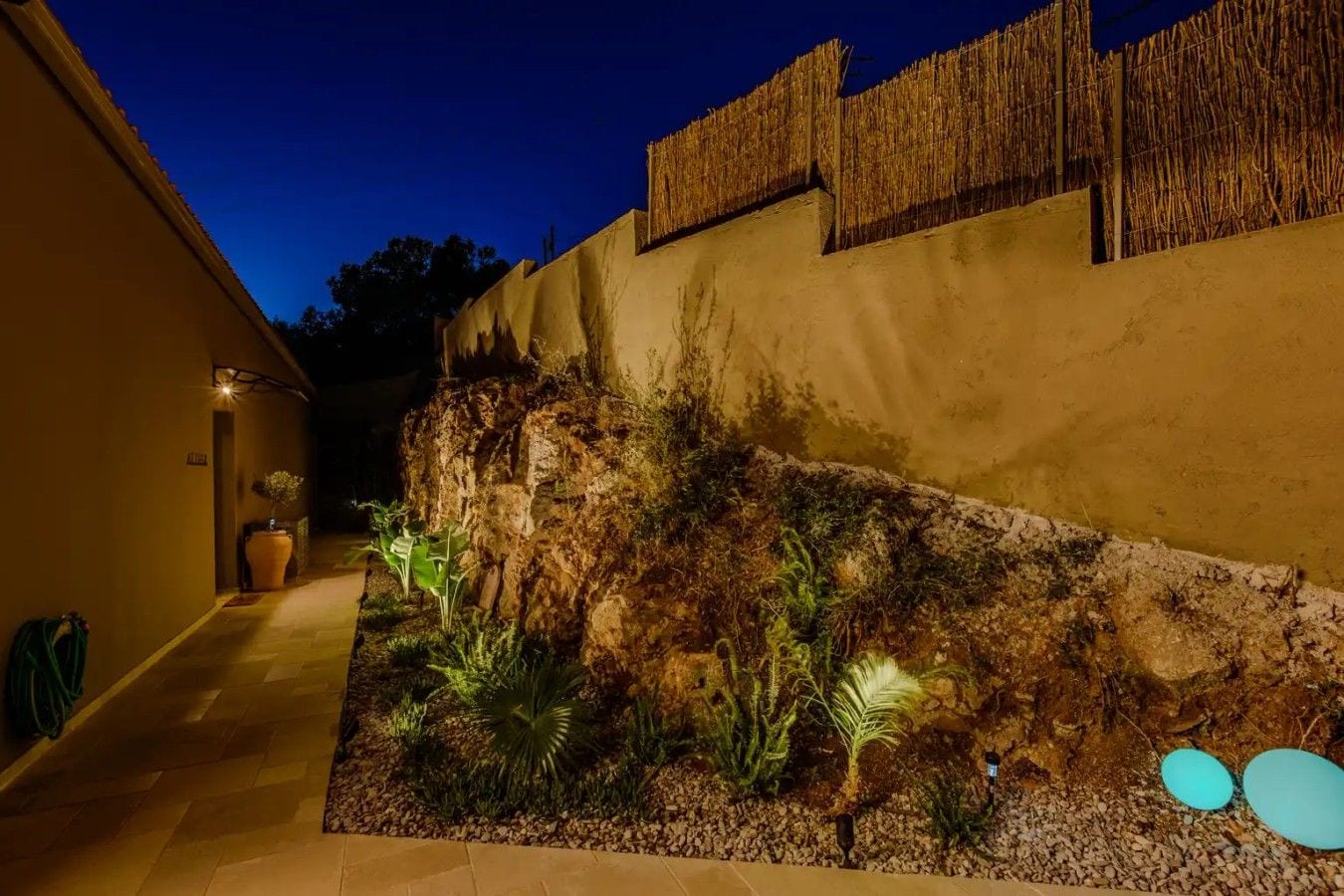 Un área de jardín ajardinada con un muro de piedra, plantas en macetas y un sendero que conduce a la entrada de un edificio.
