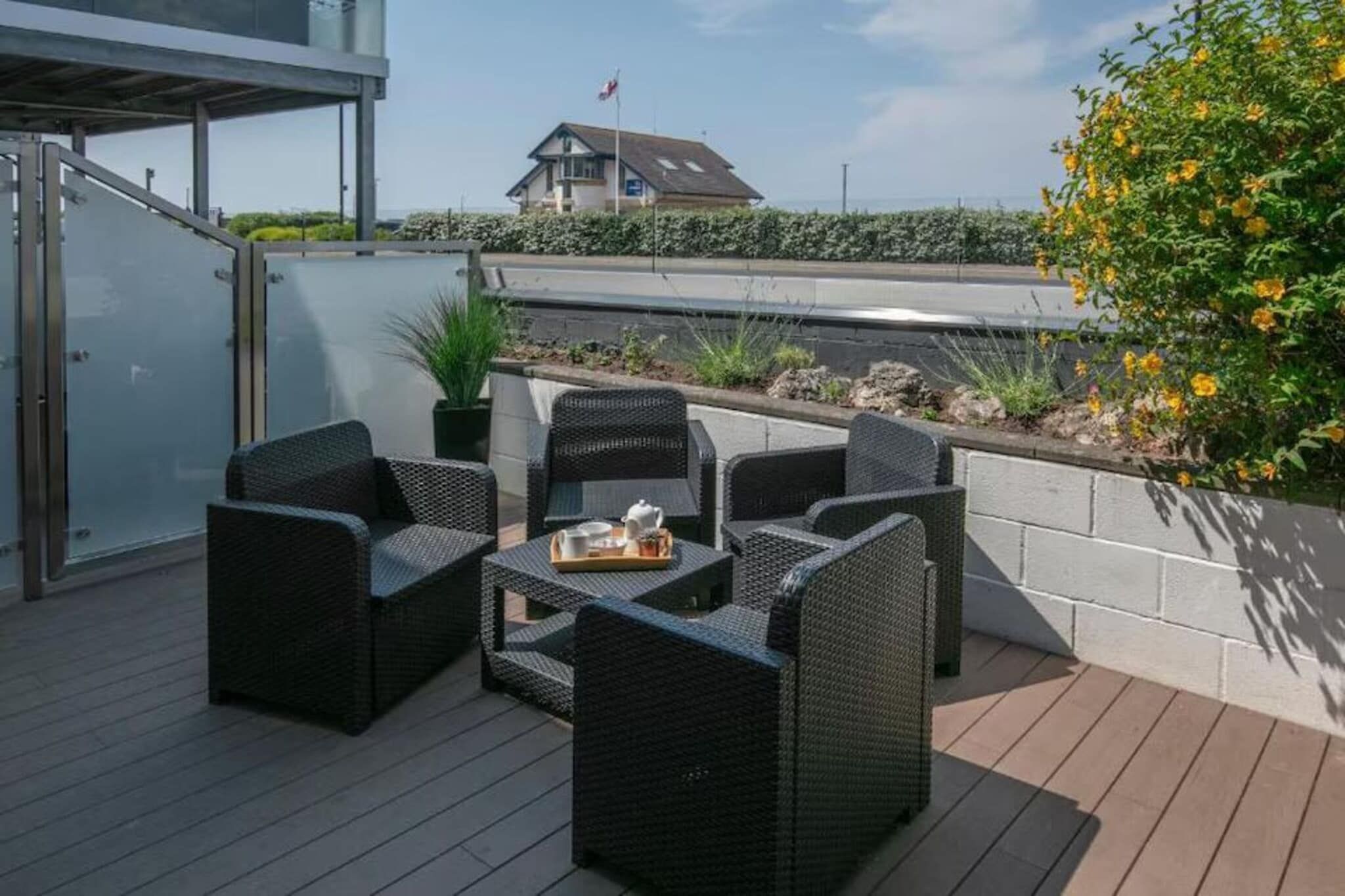 A deck with wicker furniture and a table set for dining, overlooking a landscaped area with a building in the background.