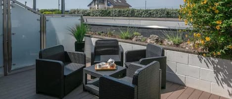 A deck with wicker furniture and a table set for dining, overlooking a landscaped area with a building in the background.