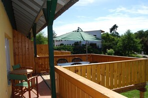 Eine Holzterrasse mit Tisch und Stühlen, einem grünen Sonnenschirm und Blick auf ein Gebäude im Hintergrund.