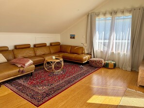Ein Wohnzimmer mit einem braunen Ledersofa, einem runden Korbtisch, einem gemusterten Teppich und einem Fenster mit durchsichtigen Vorhängen.