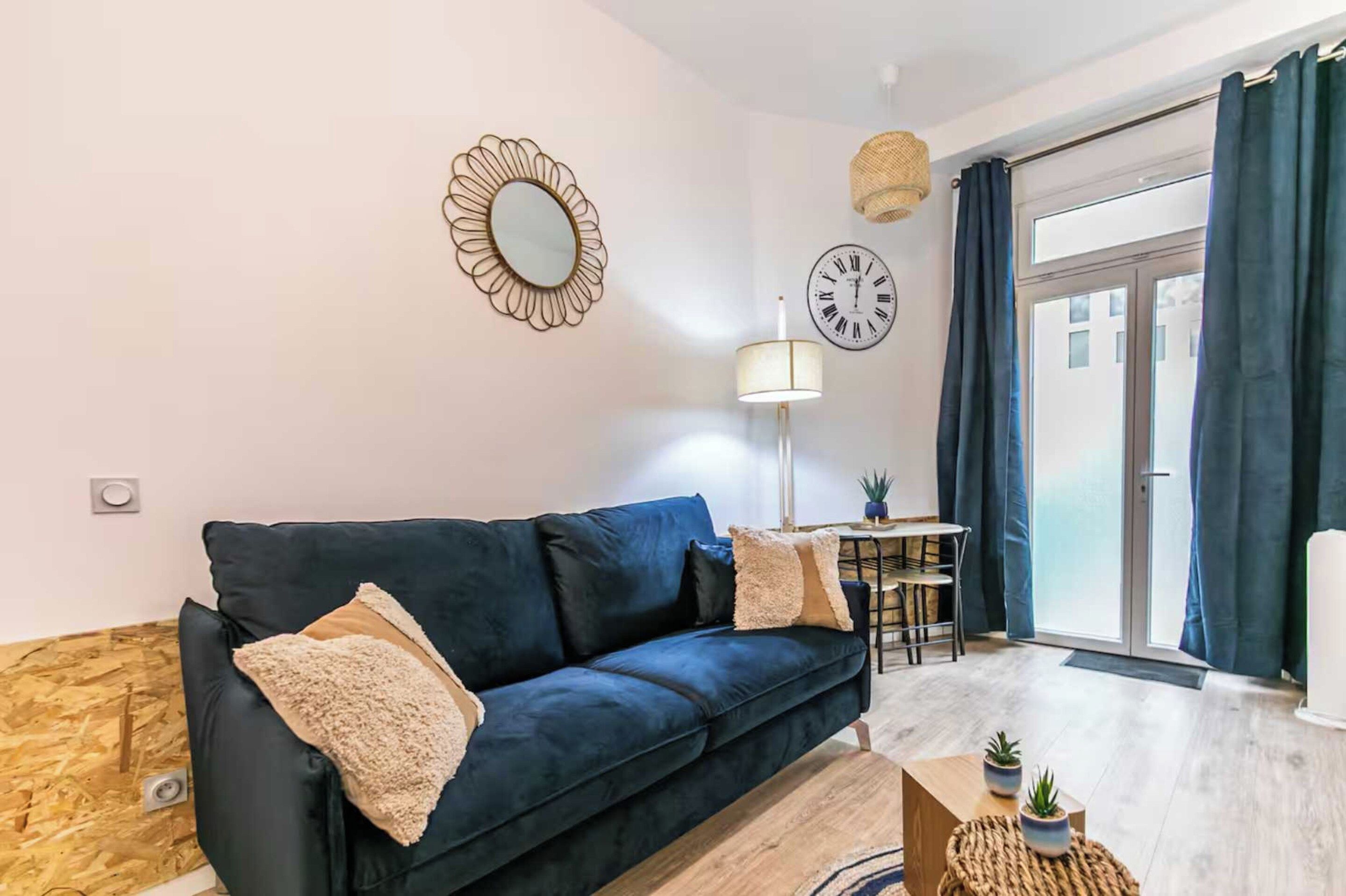 A modern living room with a dark blue sofa, a round mirror, a clock, and a dining area with a table and chairs.