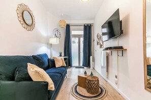 A modern living room with a dark blue sofa, a flat-screen TV mounted on the wall, a round mirror, and a small side table with a lamp. - Cosy Studio - 2P - Parmentier / Voltaire (Paris)