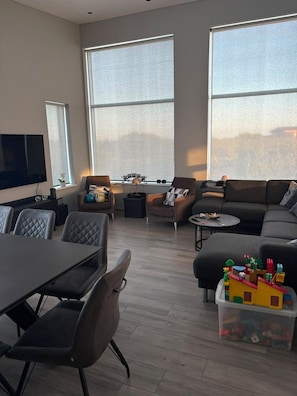 A modern living room with a dining area, large windows, a television, and a toy storage unit.