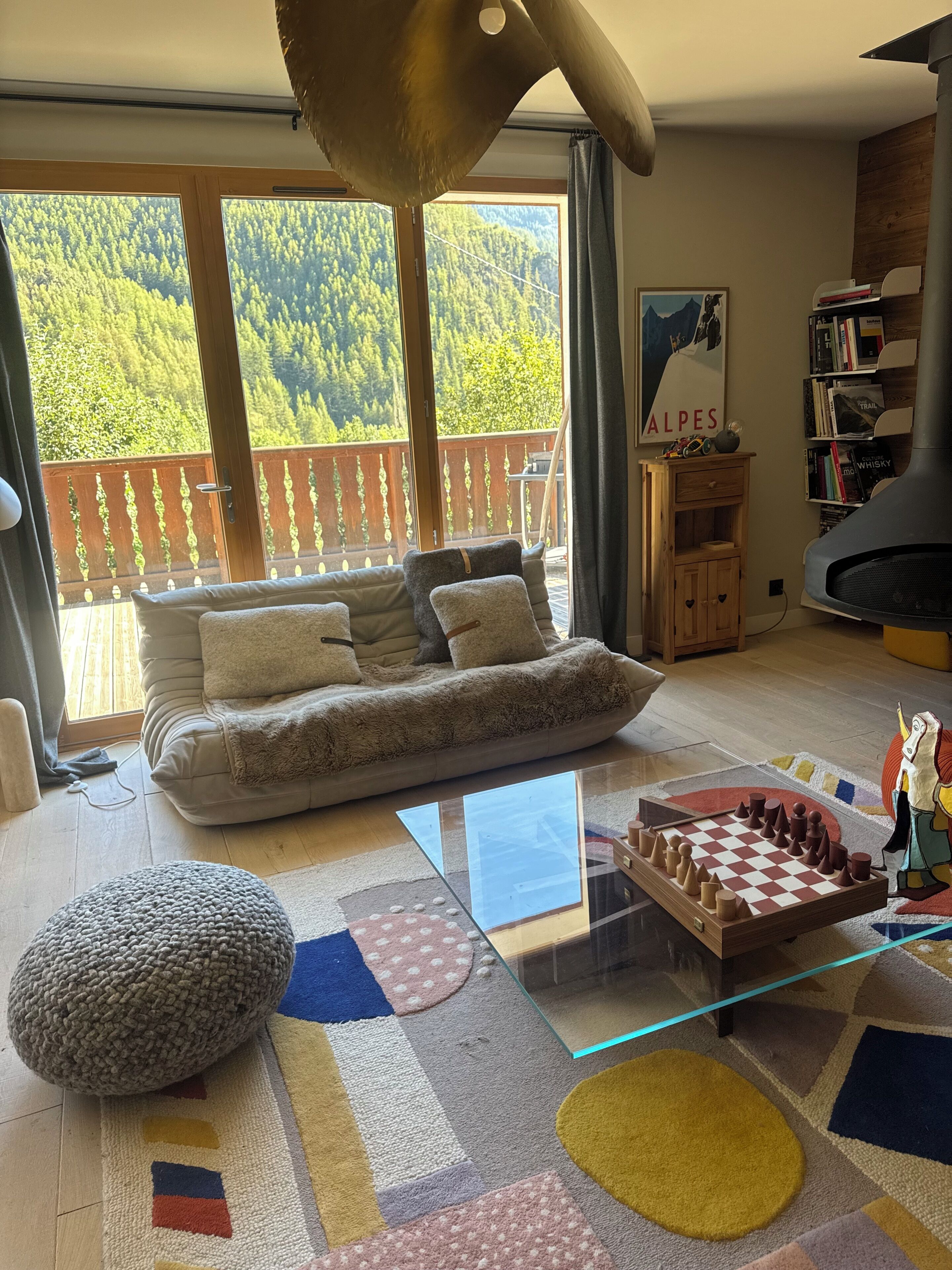 Ein Wohnzimmer mit einer Couch, einem Glastisch, einem Schachspiel, einem gestrickten Pouf und einem Bücherregal.
