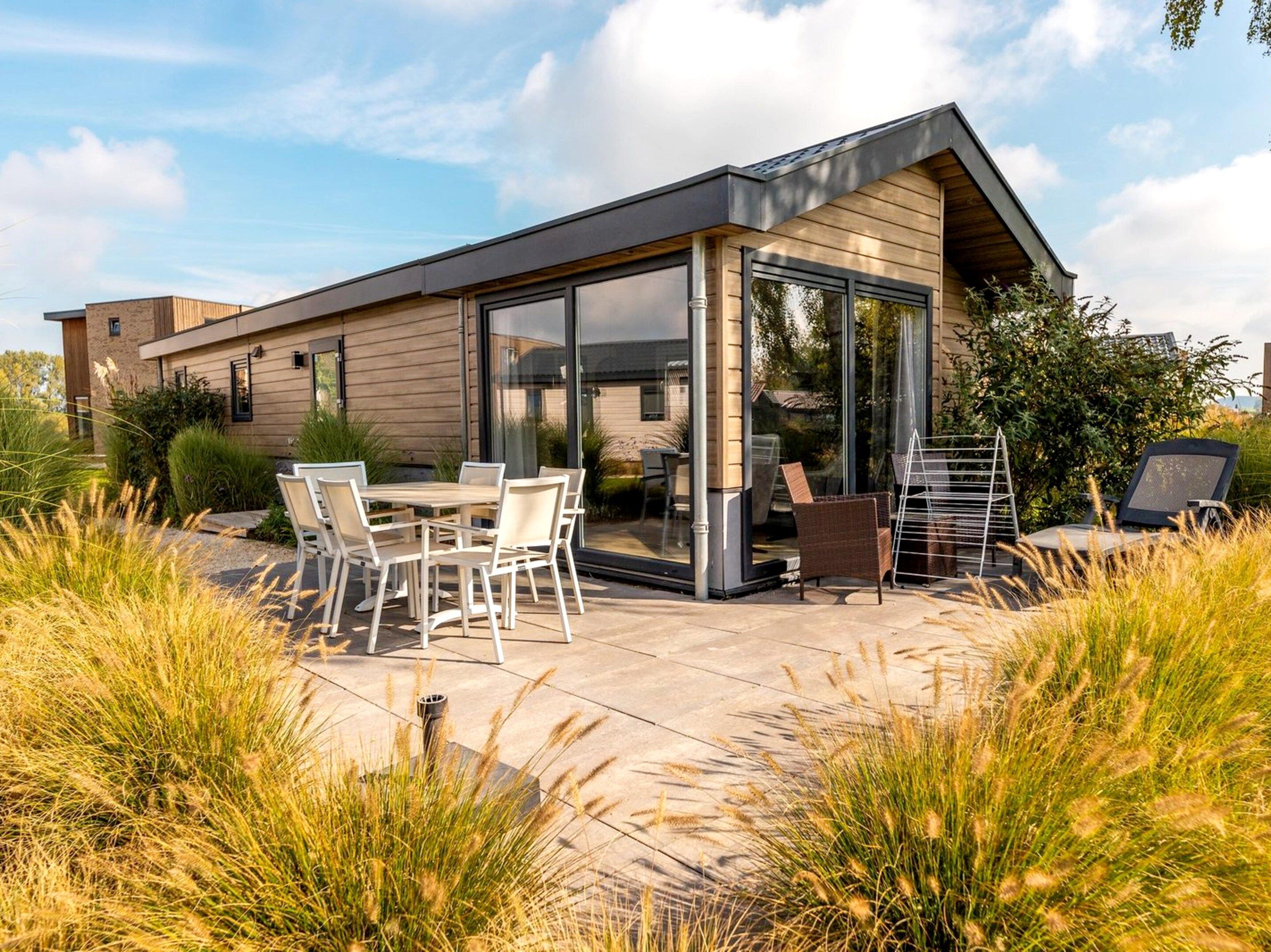 A modern house with a patio area featuring a table and chairs, surrounded by tall grasses.