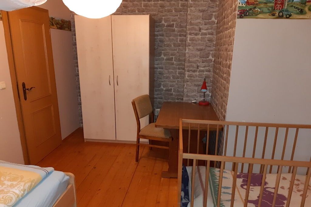 Une pièce avec un plancher en bois, un lit recouvert d’une couvre-lit à motifs, une armoire en bois, une table avec une chaise et un berceau.