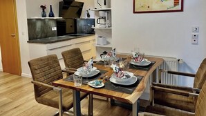 Ein Essbereich mit einem Holztisch, gedeckt für vier Personen, umgeben von Stühlen. Im Hintergrund ist eine Küche mit modernen Geräten erkennbar.