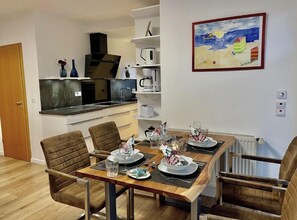 Een eetruimte met een houten tafel voor vier personen, omringd door stoelen. Achtergrond toont een keuken met moderne apparatuur.