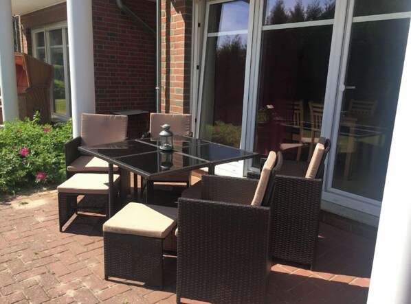 Een terras buiten met een glazen tafel en rieten stoelen.