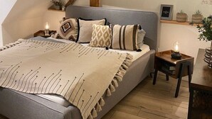 Ein Schlafzimmer mit schrägen Wänden, einem Bett mit grauem Kopfteil, Nachttischen mit Lampen und einer kleinen Pflanze auf der rechten Seite.