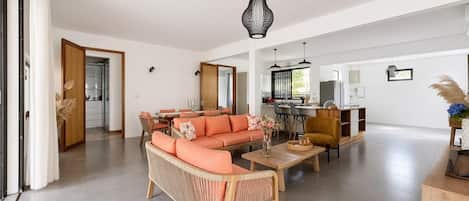 Un grand espace de vie comprenant un canapé, une table basse et une table à manger. Au fond, on aperçoit une cuisine et une porte qui donne sur une autre pièce.