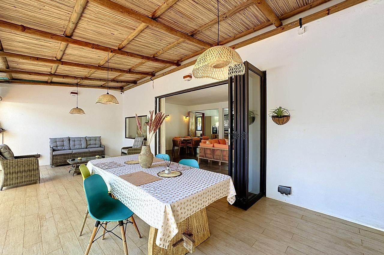 An open-plan living and dining area with a high wooden ceiling, a dining table with a patterned tablecloth, and a sofa with cushions.