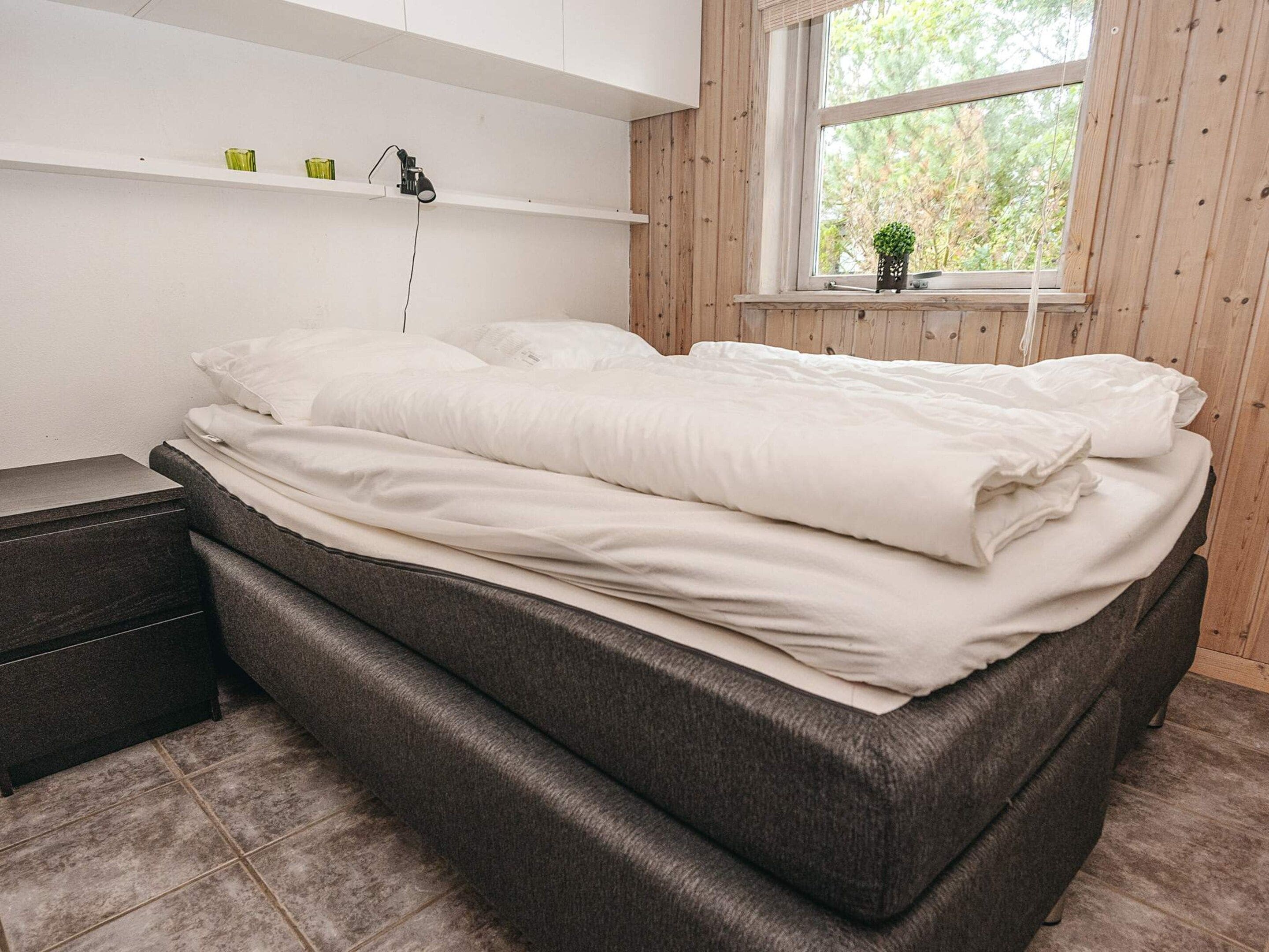 A neatly made bed with white bedding, a bedside table, a shelf with a lamp, and a window with a view of trees.