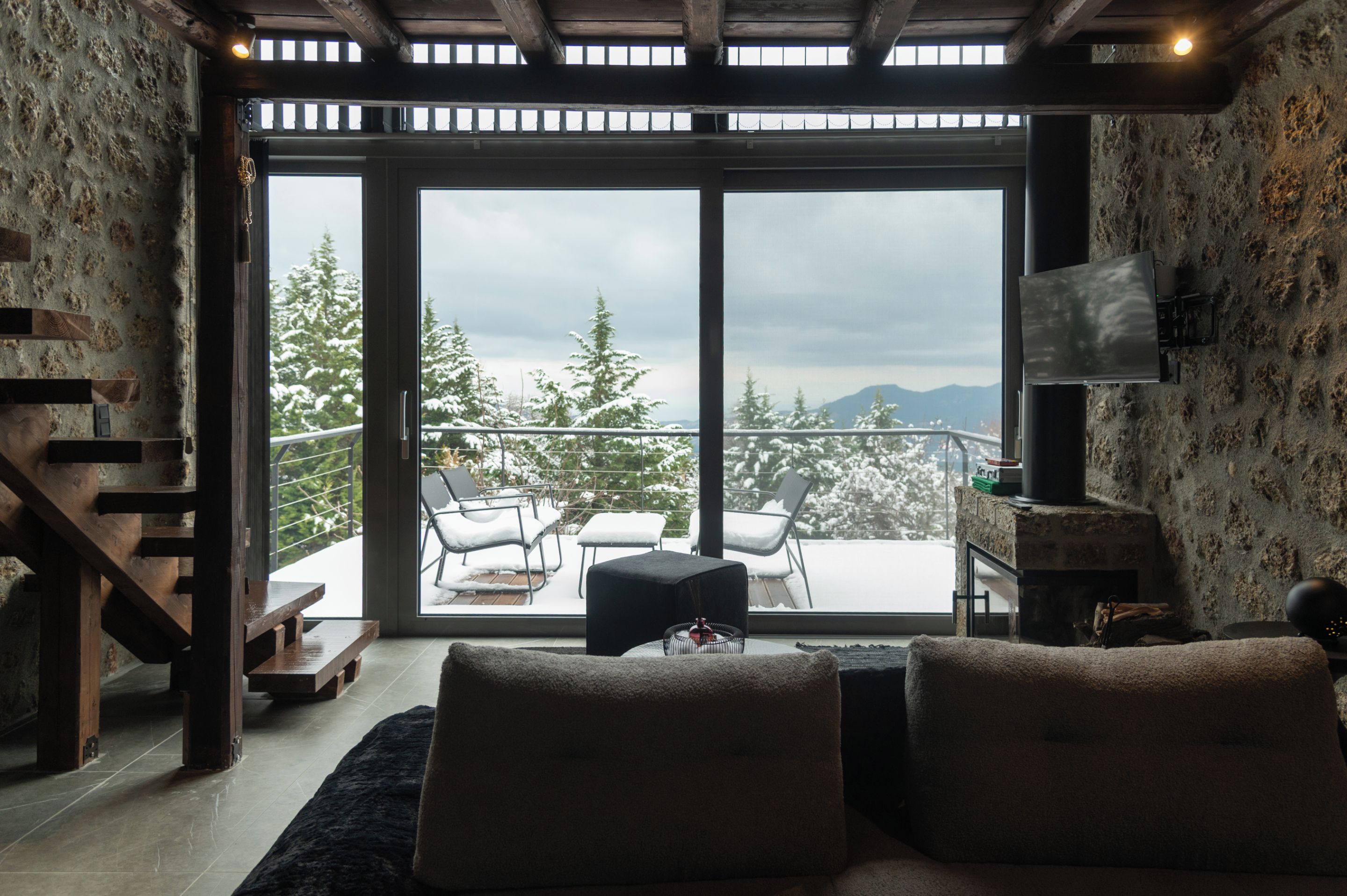 A cozy living room with a fireplace, a sofa, and a view of snow-covered trees through large windows.