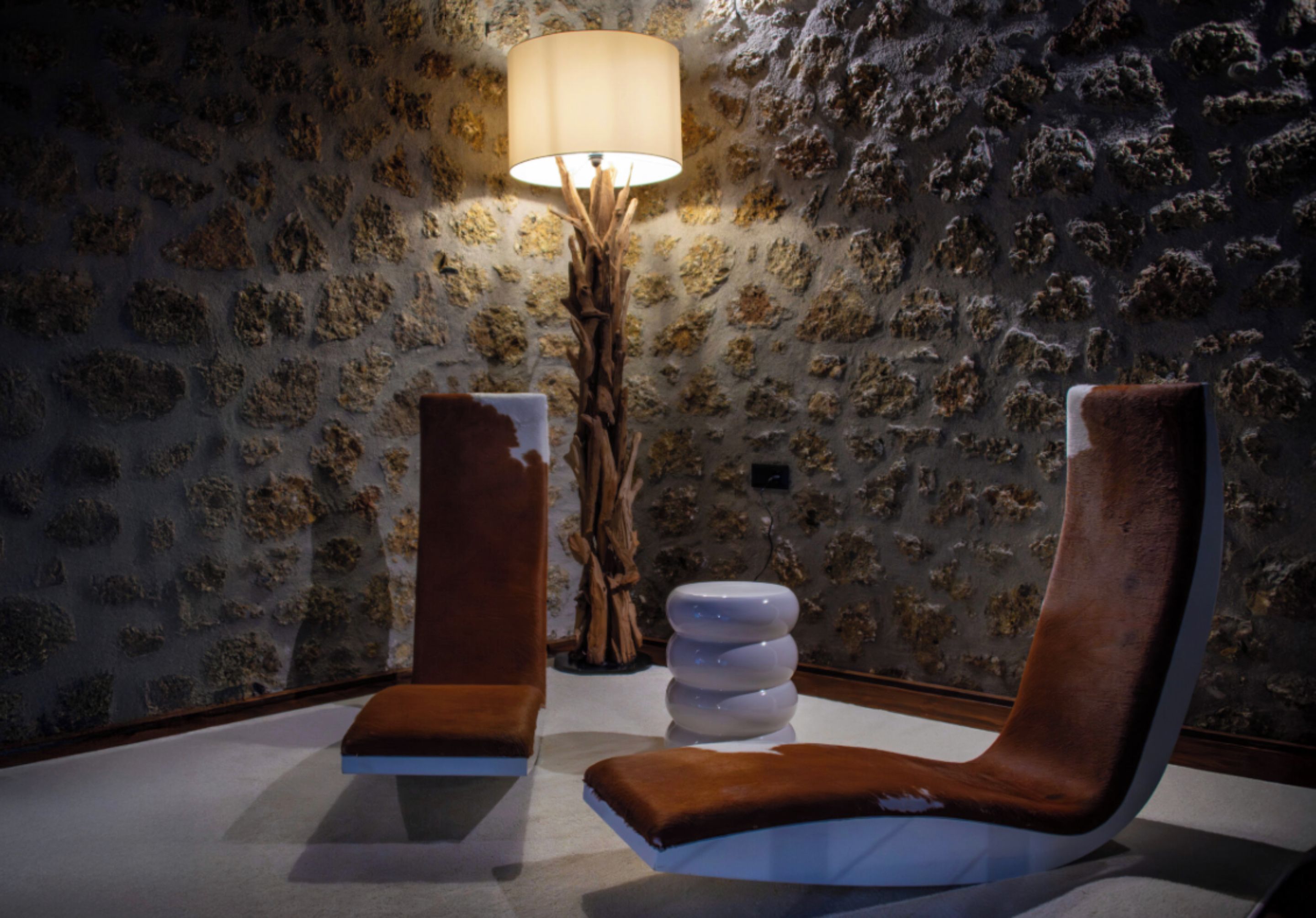 Een kamer met een stenen muur, een houten vloerlamp en twee bruine fauteuils.