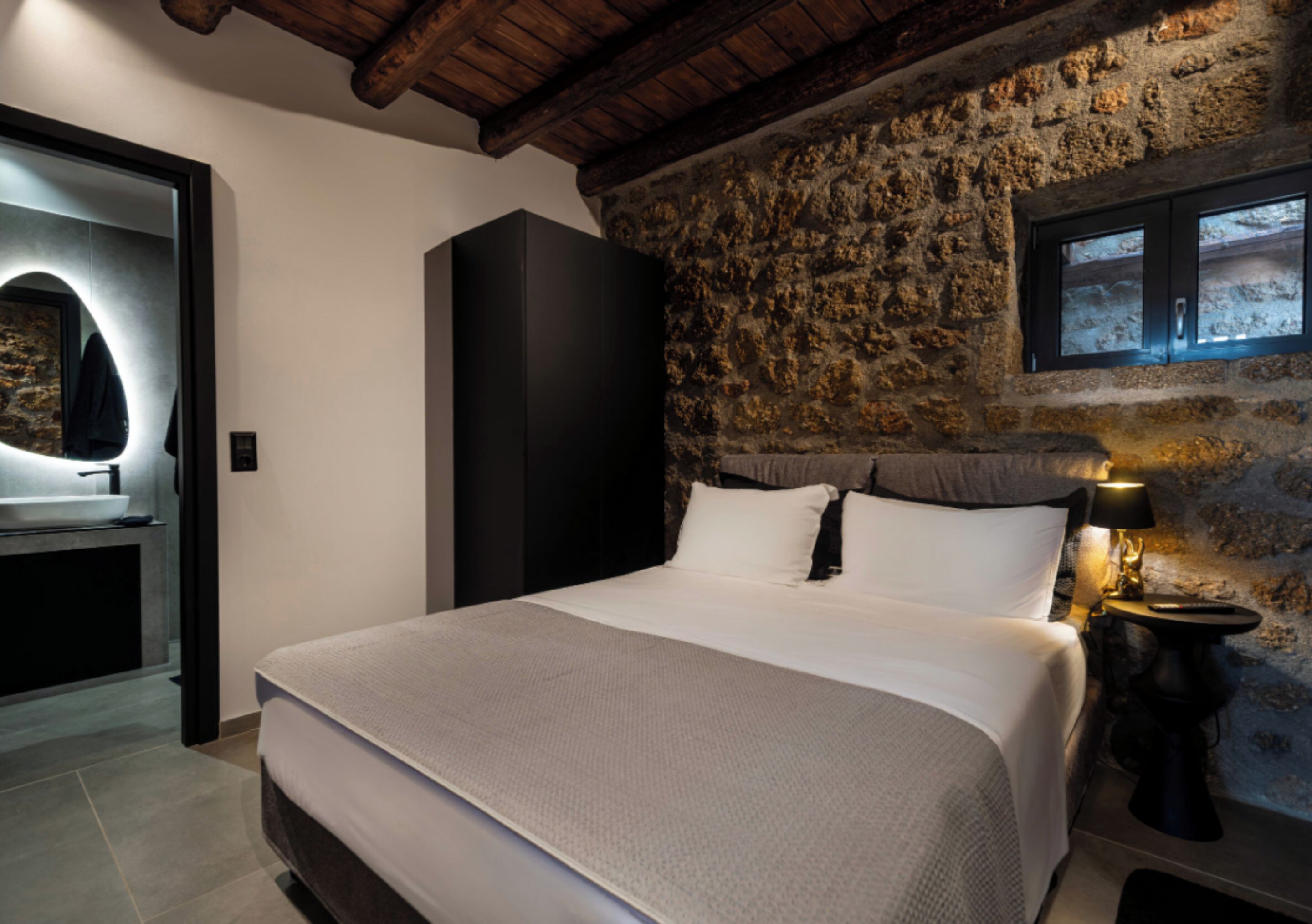Een slaapkamer met een stenen muur, een bed met witte lakens, een nachtkastje met een lamp en een spiegel met een ronde lijst.