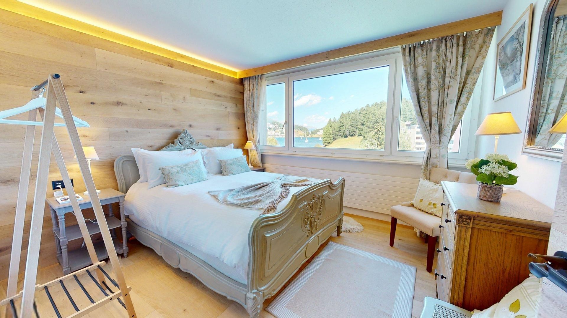 A bedroom with a wooden headboard, a bed with white linens, a bedside table with a lamp, a dresser, and a window with a view of the outside.