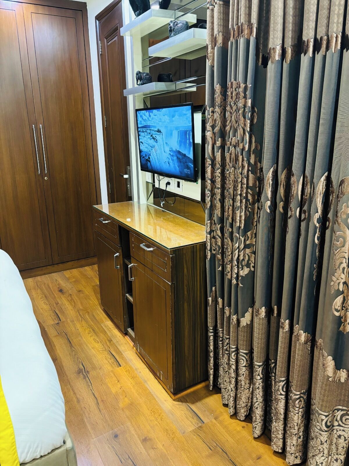 A room with a wooden wardrobe, a television on a stand, a bed with a white and yellow bedspread, and patterned curtains.