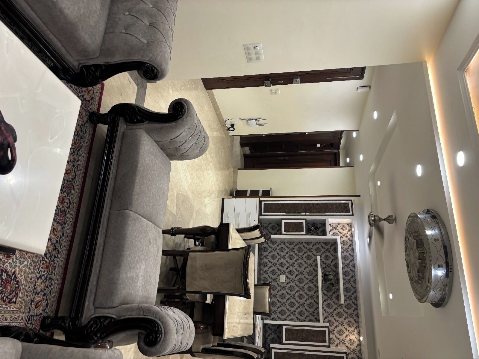 A room with a sofa, armchairs, a dining table, and a kitchen area with cabinets and a patterned backsplash.